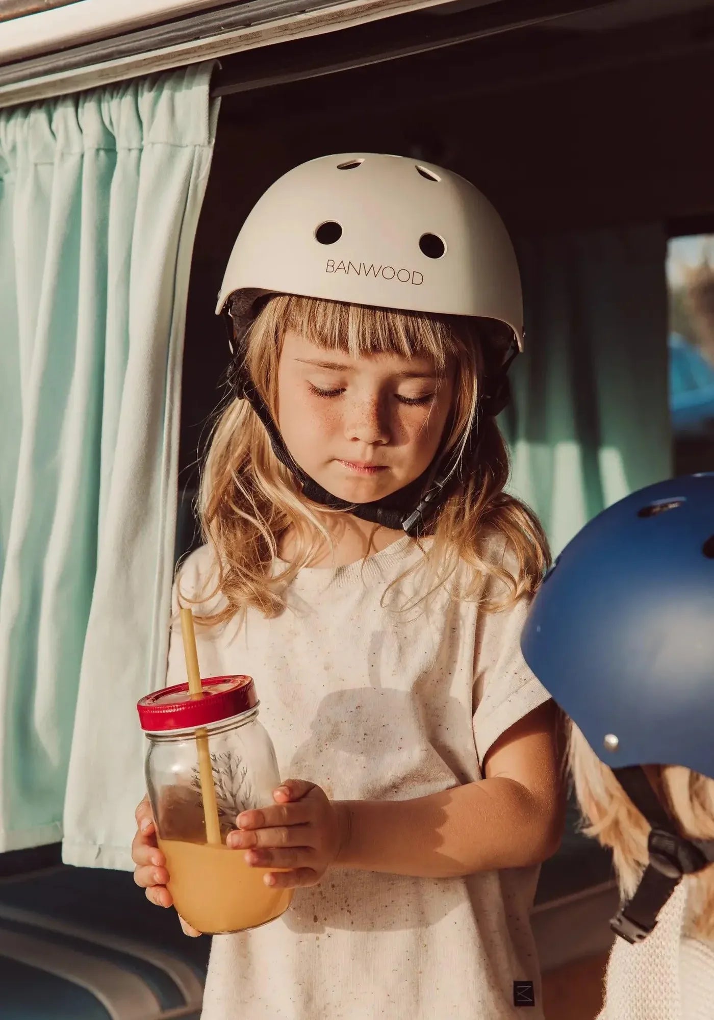 Kind mit Banwood-Helm, trinkt Saft vor Camper – skandinavischer Outdoor-Lifestyle für Kids