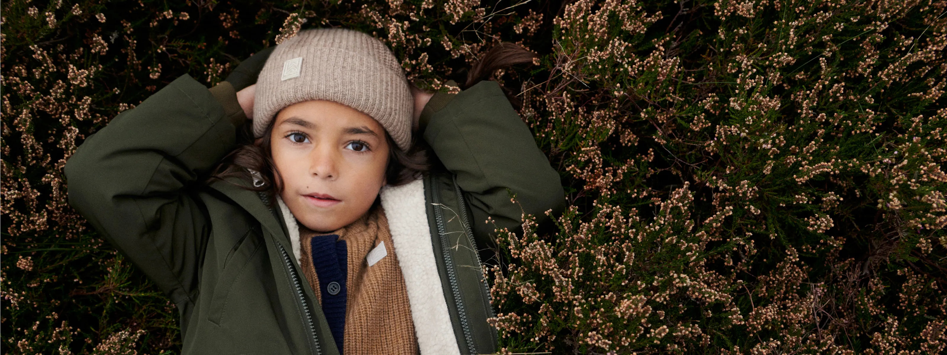Kind mit LIEWOOD Winterjacke und Strickmütze liegt in herbstlicher Heidelandschaft.